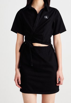 Kobieta w czarnej, krótkiej koszulce z krótkim rękawem z wiązaniem z boku oraz dopasowanej czarnej spódnicy, prosty wzór, logo Calvin Klein na koszulce.
