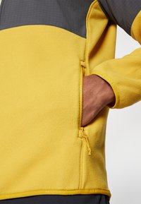 Fleece dzseki élénk sárga alappal és szürke betéttel. Zipzáros zsebet és elasztikus mandzsettákat tartalmaz, texturált felülettel.