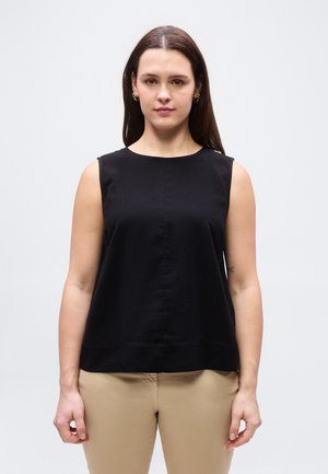 Γυναίκα με μακριά καστανά μαλλιά φοράει μαύρο αμάνικο τοπ και μπεζ παντελόνι, στέκεται μπροστά σε απλό ανοιχτό γκρι φόντο.