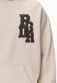 Beige hoodie med en framficka, med svarta upphöjda broderade bokstäver "PGDR" placerade vertikalt på vänster sida.