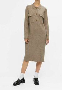 Robe en maille côtelée beige, longueur genou, avec col, boutons marron, manches longues, deux poches poitrine ; modèle portant des chaussettes blanches et des mocassins noirs.