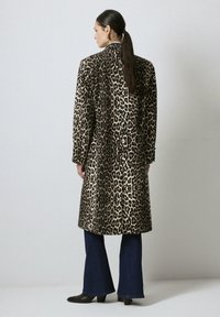 Femme avec de longs cheveux attachés portant un manteau en imprimé léopard de longueur genou, un jean flare noir et des bottes à talons noires, de dos sur fond uni.