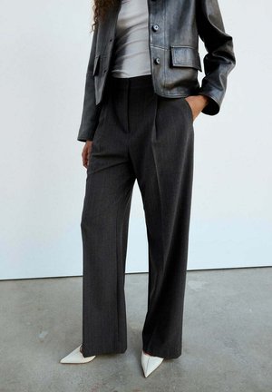 Donna che indossa pantaloni scuri a gamba larga, scarpe bianche con punta, un top bianco e una giacca corta in pelle nera con tasche e bottoni.