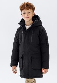 Parka noire rembourrée avec capuche, dotée de poches zippées sur les côtés et d'un écusson sur la manche gauche. Texture lisse et matériau chaud.