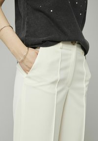 Pantalons blancs ajustés en tissu lisse, avec des plis avant et des poches latérales, associés à une chemise gris foncé avec des embellissements décoratifs.