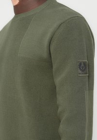 Olive green knitted sweater featuring textured accents and a patch logo on the left sleeve, showcasing a crew neckline and ribbed cuffs.