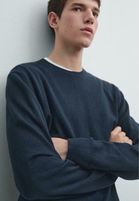 Granatowy dzianinowy sweter z okrągłym dekoltem i długimi rękawami, o fakturowanym wykończeniu i ściągaczach na mankietach. Modelka ma skrzyżowane ramiona.