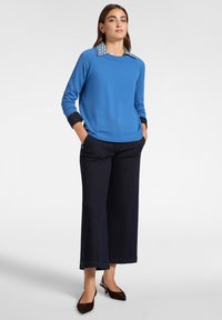 Maglione blu a maglia con colletto decorato, abbinato a pantaloni ampi scuri e tacchi a fascia marroni. Texture liscia, vestibilità rilassata.