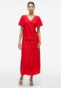 Sarkans, īsām piedurknēm, V veida kakla kleita ar plūstošu svārku. Izgatavota no viegla auduma, ar pūkainām piedurknēm un sakrautu jostasvietu, kas piešķir apjomu.