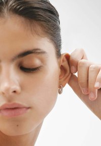 Pendiente de aro de plata con un diseño de eslabones de cadena texturizados, llevado en una oreja. El modelo tiene un maquillaje neutro y piel suave y clara.
