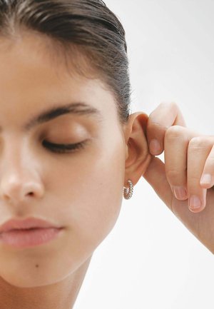 Pendiente de aro de plata con un diseño de eslabones de cadena texturizados, llevado en una oreja. El modelo tiene un maquillaje neutro y piel suave y clara.