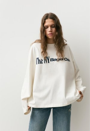 Junge Frau mit langen Haaren, die ein weißes, übergroßes Sweatshirt mit der Aufschrift "The NY Bagel Co." und blaue Jeans trägt, vor einem einfarbigen Hintergrund.