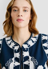 Marinblå broderad jacka med vita blommönster, med rund halsringning och knäppning. Mjuk textur med intrikata detaljer.