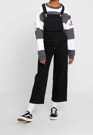 Sorte denim overalls med brede ben, der har en frontlomme og justerbare stropper. Sat sammen med en grå og hvid stribet sweatshirt og sorte sneakers.