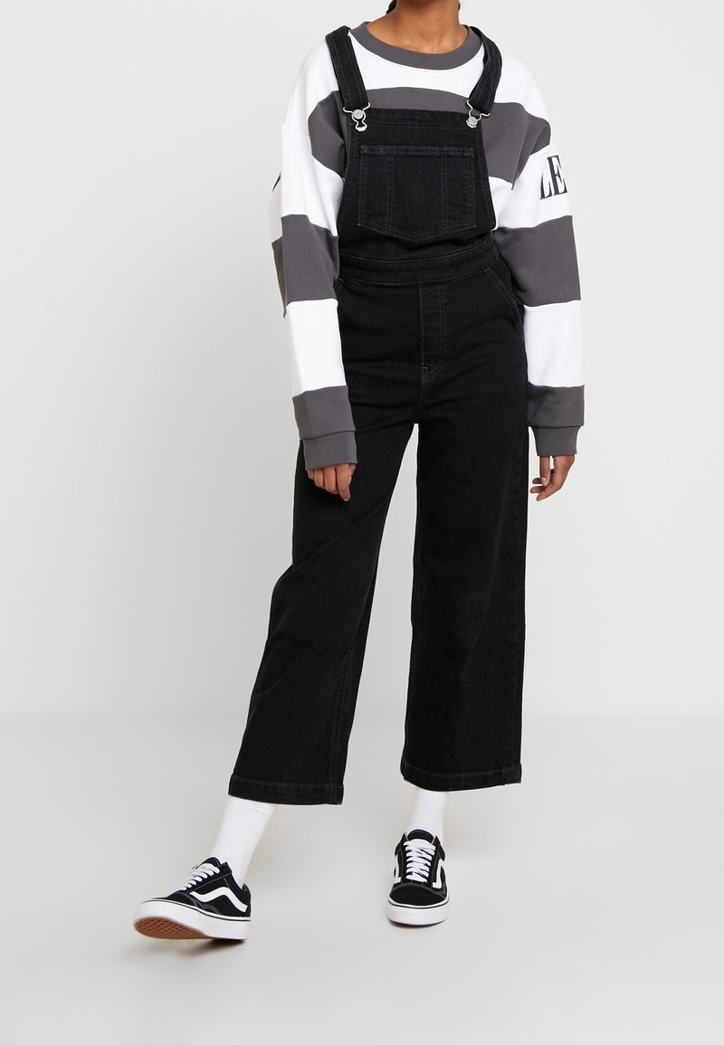 Salopette en denim noir à jambes larges, dotée d'une poche avant et de bretelles ajustables. Associée à un sweat-shirt rayé gris et blanc et à des baskets noires.