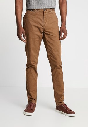 Man wearing slim-fit brown pants and maroon sneakers, standing with hands relaxed by sides against plain background.