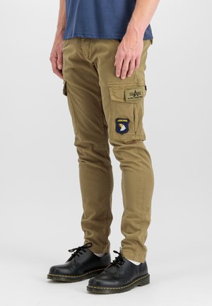 Mann trägt khakifarbene Cargohosen mit Airborne- und Alpha Industries-Aufnähern, schwarze Schnürschuhe und ein blaues Hemd, steht vor weißem Hintergrund.