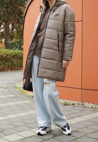 Padded olive green long coat with a hood, black logo, two side zip pockets, paired with light blue ripped jeans and black sneakers.