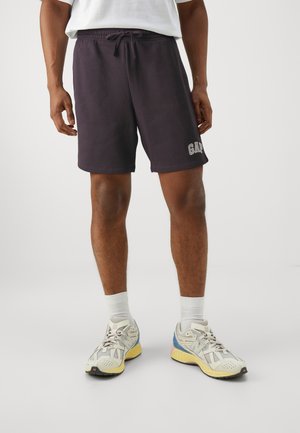 Mann trägt schwarze GAP-Shorts, weiße Socken und mehrfarbige New Balance-Turnschuhe vor einem einfarbigen Hintergrund.