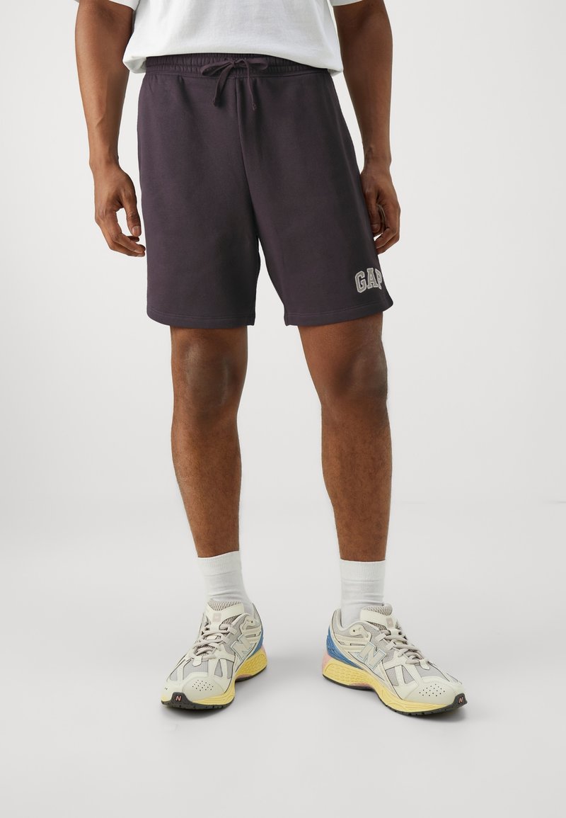 Homme portant un short noir GAP, des chaussettes blanches et des baskets multicolores New Balance sur un fond uni.
