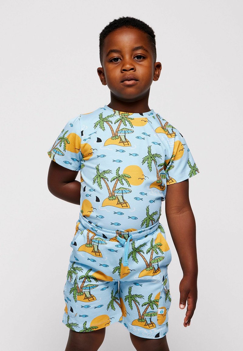 Jeune garçon debout, un bras derrière le dos, portant une chemise et un short assortis bleu clair avec un motif de palmier, soleil, poisson et île.