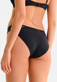 Ensemble de lingerie noire en tissu lisse et opaque avec une bordure en dentelle au niveau de la taille. Le design comprend une coupe mi-haute et des bords sans couture.