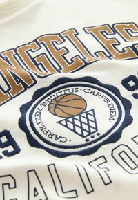 Texto de Los Ángeles en marrón y azul marino sobre una tela blanca, con un emblema de baloncesto y aro, incluyendo la frase en latín 'Carpe Diem'.