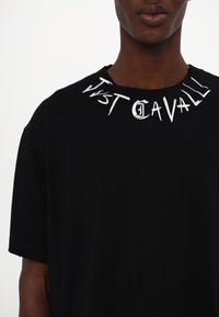 Black t-shirt made of cotton with a round neckline. Features white printed text "Just Cavalli" around the collar, with a distressed edge.