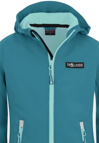 Tägliche Softshelljacke in Teal mit einer hellblauen Innenseite, Frontreißverschluss und zwei Seitentaschen. Mit einem schwarzen Logopatch auf der Brust.