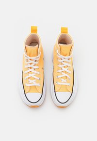 Jasnożółte sneakersy typu high-top wykonane z płótna z białymi sznurowadłami, czarnymi akcentami i gumową podeszwą. Posiadają tylną pętelkę ułatwiającą zakładanie.