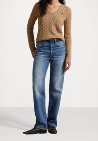 Femme portant un pull col en V en maille torsadée beige, un jean bleu droit avec une ceinture noire, et des bottes à bout pointu marron foncé, debout.