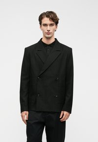 Νεαρός άνδρας με σπαστά μαλλιά, φορώντας μαύρο σακάκι με διπλό κούμπωμα και ρίγες πάνω από μαύρο πουκάμισο και μαύρα παντελόνια, μπροστά σε ουδέτερο φόντο.