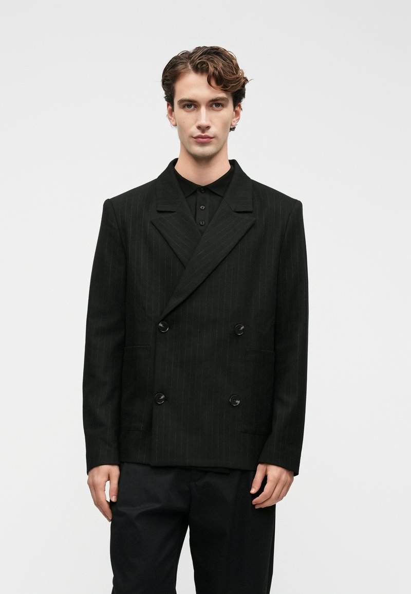 Νεαρός άνδρας με σπαστά μαλλιά, φορώντας μαύρο σακάκι με διπλό κούμπωμα και ρίγες πάνω από μαύρο πουκάμισο και μαύρα παντελόνια, μπροστά σε ουδέτερο φόντο.