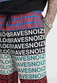 Bunte Shorts mit einem sich wiederholenden Textmuster von "BRAVESNOIZE" in Rot, Weiß, Schwarz und Teal auf einem glatten Stoff. Elastischer Bund mit Kordelzug.