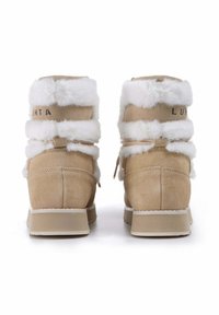 Vaahto beige-nahasta valmistetut nilkkurit, joissa on valkoinen tekoturkistrimmi, tasapohja ja lenkki pukeutumisen helpottamiseksi. Brändin nimi näkyy takana.