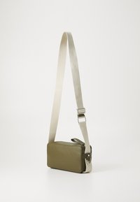 Green leather crossbody bag with a rectangular shape, cream fabric strap, silver hardware, and a zip closure. Smooth texture, minimalist design.