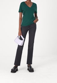 Grön v-ringad t-shirt, svarta raka jeans och svarta plattformsskor. Håller en liten vit handväska med lila accenter.