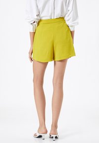 Shorts de color verde lima con un acabado texturizado, con una cintura alta y dobladillo acampanado. Combinados con sandalias de tacón abiertas en la parte delantera y blancas.