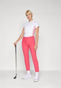 Γυναίκα φορώντας λευκό πόλο μπλουζάκι και ροζ παντελόνι, κρατώντας ένα γκολφ, στέκεται σε λευκό φόντο.