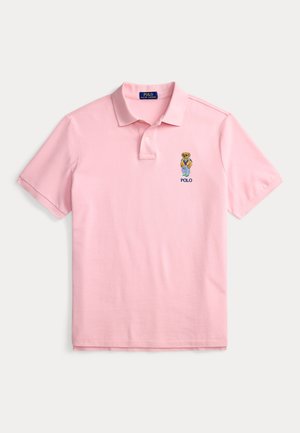Tricou polo roz deschis cu mâneci scurte, cu un ursuleț brodat mic care poartă haine casual și textul „POLO” pe piept, în partea stângă.