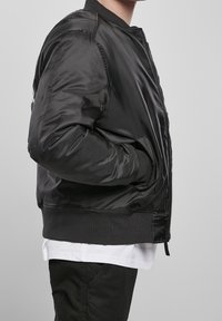 Giubbotto bomber nero con finitura lucida, polsini e colletto a coste. Presenta una zip frontale e tasche laterali. Indossato sopra una camicia bianca.
