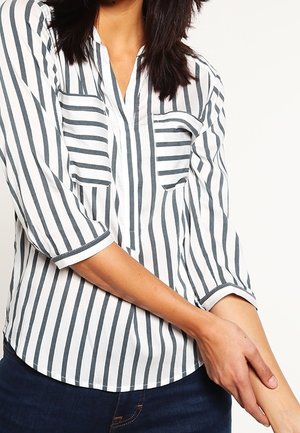 Femme portant une chemise blanche à rayures verticales noires et manches trois-quarts, tenant son avant-bras gauche avec sa main droite.