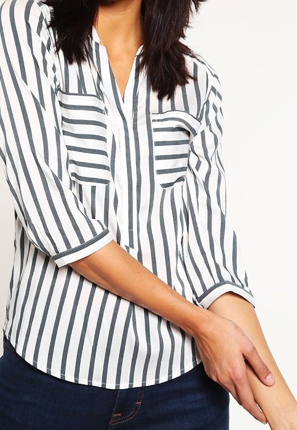 Femme portant une chemise blanche à rayures verticales noires et manches trois-quarts, tenant son avant-bras gauche avec sa main droite.