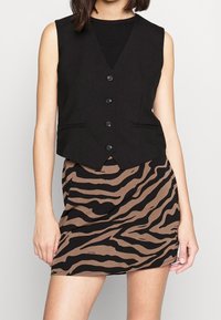 Schwarzes ärmelloses Weste mit fünf Knöpfen und zwei Taschen, kombiniert mit einem braunen Rock mit einem schwarzen Zebra-Streifen-Muster.