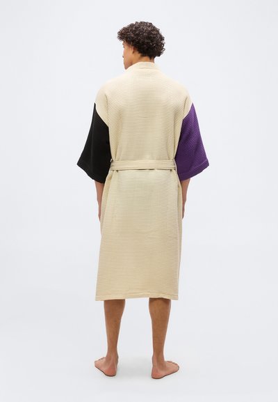 Άτομο με σγουρά μαλλιά που φορά ρόμπα μήκους μέχρι το γόνατο, μπεζ με υφή βάφλας, με μαύρο αριστερό μανίκι και μωβ δεξί μανίκι, όρθιο ξυπόλυτο με την πλάτη προς τα έξω.