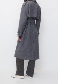 Abrigo de lana gris con cintura ceñida por un cinturón, detalle de solapa en la parte posterior y puños abotonados, llevado sobre pantalones oscuros y calzado crema.