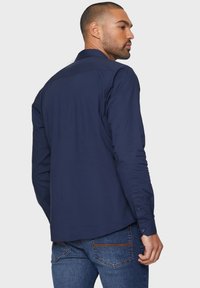 Navy lange mouwen shirt, met knoopmanchetten, gladde katoenen stof, getailleerde pasvorm, ronde zoom en rug yoke details. Draagt met blauwe spijkerbroek.
