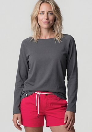 Frau mit schulterlangem blondem Haar, die ein dunkelgraues Langarmshirt und leuchtend pinke Shorts mit weißem Kordelzug trägt.