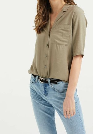 Kvinde iført en lys olivengrøn kortærmet skjorte med knapper, stoppet ned i lyseblå jeans med sort bælte og to ringe på hendes højre hånd.