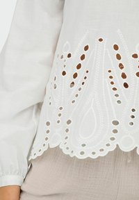 Weißes, leichtes Oberteil mit filigraner Cut-Out-Stickerei am Saum und gewelltem Rand; lange Ärmel mit gerafften Manschetten.